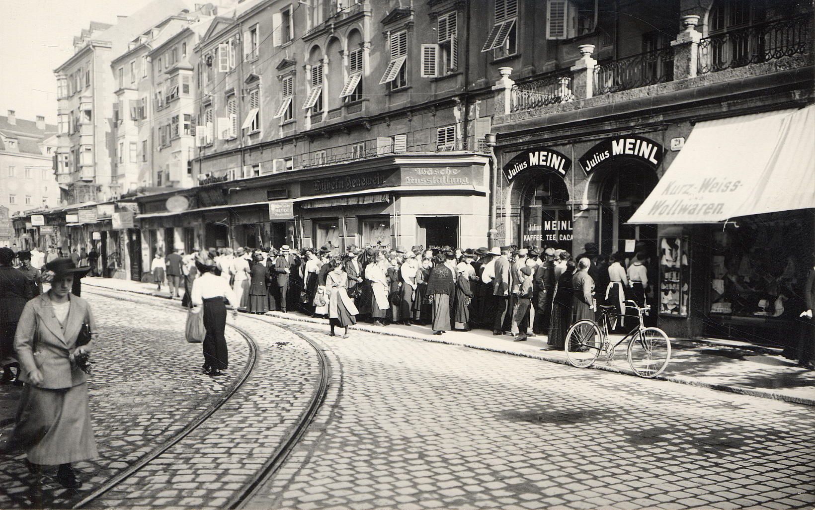 Dichtes Gedränge am Wegesrand. Kein untypischer Anblick, wenn es um die Lebensmittelausgaben in Innsbruck geht. Eine Gruppe Menschen stellt sich vor dem Meinl-Geschäft am Marktgraben an. Aufgenommen wurde dieses Foto ungefähr 1920.
